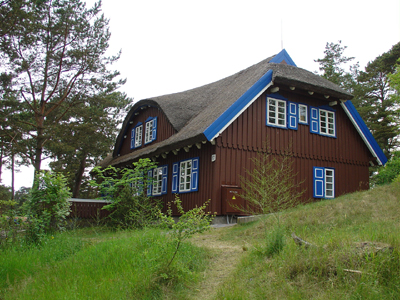 Wanderreise auf der Kurische Nehrung Ein Wanderweg in der Natur führt an einem alten Holzhaus mit blauen Akzenten vorbei