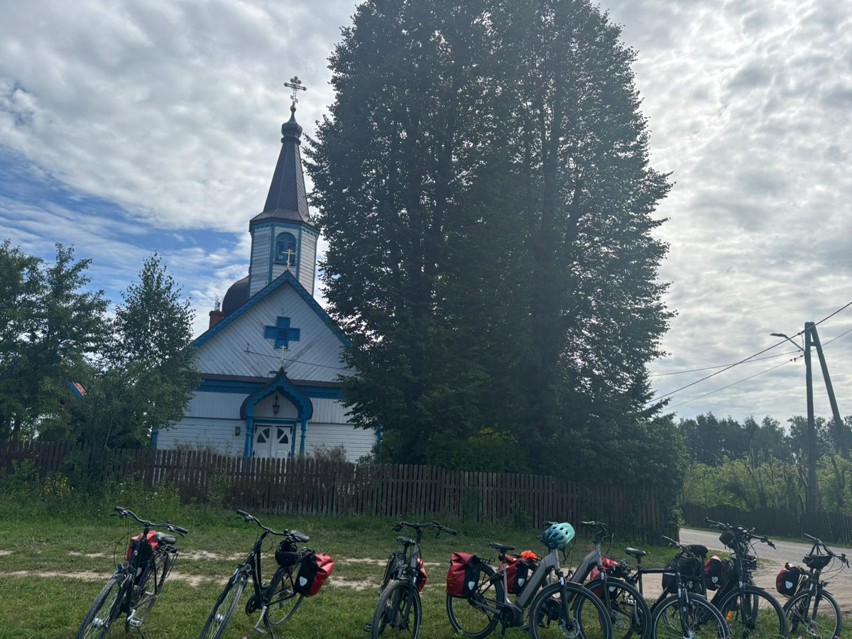 Aufnahme von der Kirche in Wojnowo mit blauen Akzenten rechts steht ein hoher Baum. Im Vordergrund stehen Fahrräder