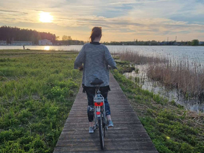 Natur und Kultur in Nordostpolen Eine Frau fährt mit dem Fahrrad auf einem Steg zwischen eine See und Gräsern bei Sonnenuntergang