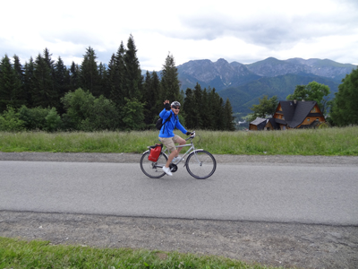 Radtour Dunajec Ein Mann fährt auf einem Radweg und winkt in die Kamera. Im Hintergrund sind Berge, Wälder und ein Holzhaus
