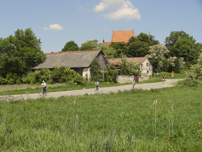 Nida-Radweg Aufnahme von Nida-Radweg mit 4 Fahrradfahrern drauf und dahinter stehen mehrere Häuser und Bäume