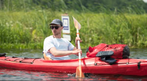 Ein Mann mit Cap und Sonnenbrille sitzt in einem Kanu und paddelt über einen See. Im Hintergrund sind Gräser