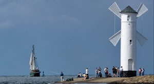 Ein weißer Leuchtturm mit Menschen davor und rechts daneben fährt eine Boot auf einem See