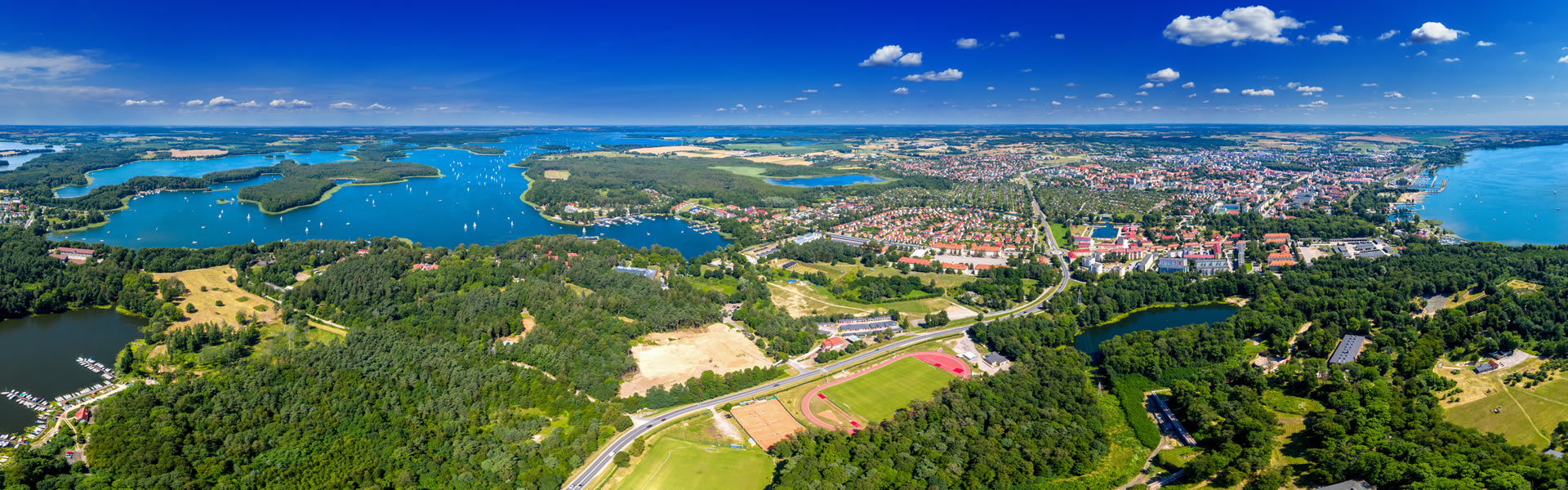 Eine weite Luftaufnahme einer Seenlandschaft mit zahlreichen blauen Seen, grünen Wäldern, Feldern und einer kleinen Stadt unter einem blauen Himmel mit vereinzelten weißen Wolken.